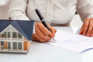 Person signs loan paperwork next to plastic model of a house.