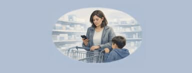 A woman stands with her child by a shopping cart in a store aisle, looking at her phone.