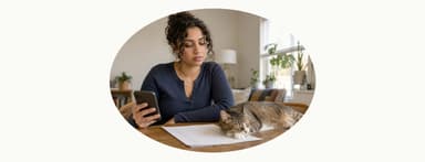 Woman looks at her phone at a table while a cat sleeps on her papers.
