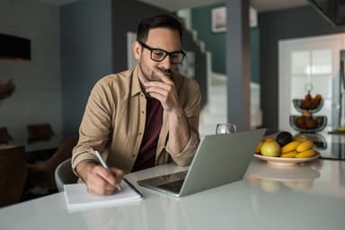 Man writes in a notebook while looking at a laptop.